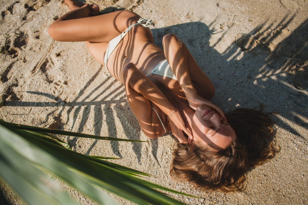 young sexy woman laying on sand beach under palm tree leaf, artistic shadow pattern on body, white bikini swim suit, tanned skin, slim body